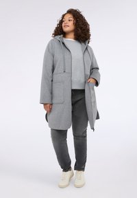 Cappotto lungo grigio con cappuccio e vestibilità rilassata, ampie tasche frontali, abbinato a una felpa grigia e jeans grigio scuro, completato da sneakers bianche.