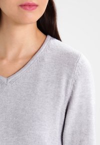 Ljust grå v-ringad tröja med en slät textur, med subtila sömnadsdetaljer längs halsringningen och axlarna.