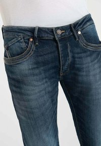 Vaqueros de denim azul oscuro con un diseño clásico de cinco bolsillos, herrajes en tono cobre y costuras en contraste. Corte slim con un ligero desgastado en los muslos.