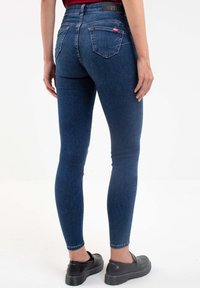 Smala jeans i mörkblå denim, med midjehöjd, klassiska bakfickor och en åtsittande siluett. Svarta slip-on skor synliga.