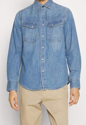 Man draagt een blauwe denim blouse met lange mouwen, knopen en twee voorzakken met kleppen en een beige broek tegen een effen achtergrond.