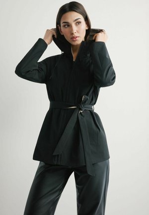 Kobieta poprawiająca kaptur długiej czarnej kurtki z paskiem, ubrana w czarne spodnie, stojąca na tle jednolitego jasnego tła.