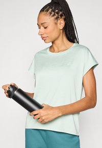 Ανοιχτόπρασινο πουκάμισο κοντών μανικιών, με ομαλή υφή, άνετη εφαρμογή. Μαύρο μπουκάλι νερού με καπάκι που στρίβει κρατιέται στο χέρι. Φοράει τιρκουάζ παντελόνι.