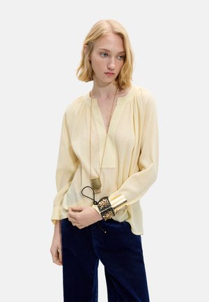 Blondynka ubrana w luźną bladożółtą bluzkę, ciemnoniebieskie spodnie, długi naszyjnik z zawieszką oraz liczne duże złote i czarne bransoletki.