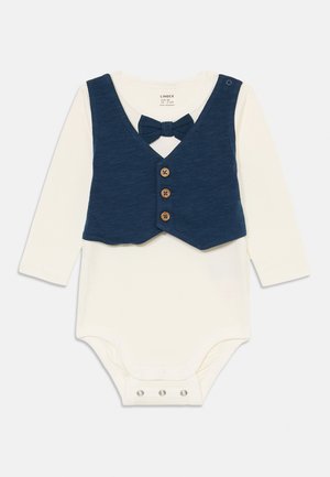 Cremfarbener Babyschlafi mit langen Ärmeln und angenähter dunkelblauer Weste, drei vorderen Holzknöpfen und passender dunkelblauer Fliege am Halsauschnitt.