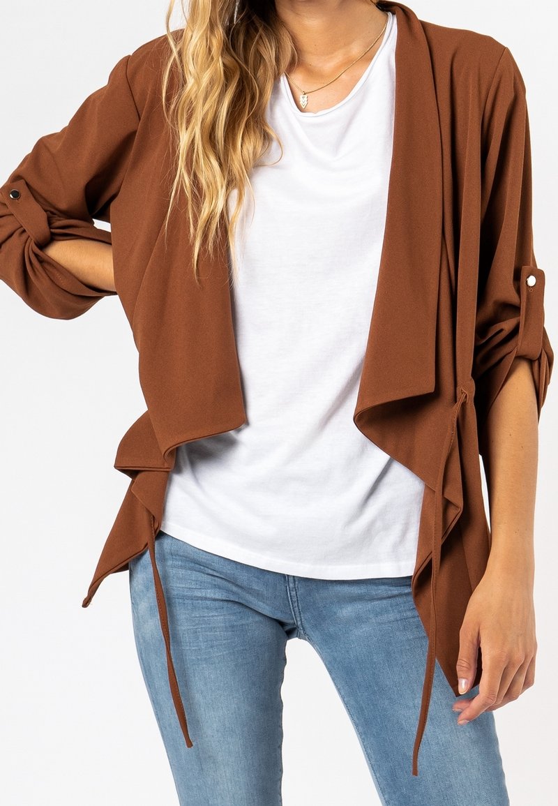 Veste légère, ouverte à l'avant, brun avec un design drapé et des fermetures à nouer. Comprend des manches roulées avec des accents de bouton. Portée par-dessus une chemise blanche.