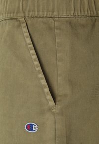 Pantalones cortos de algodón verde oliva con bolsillo lateral, cintura elástica y un parche del logo de Champion en rojo, blanco y azul. Detalle de tela texturizada visible.