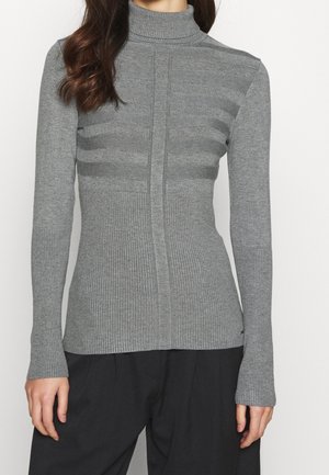 Femme portant un pull col roulé gris ajusté en maille côtelée avec de subtiles rayures horizontales et un pantalon noir ample, visible du cou jusqu'aux hanches.