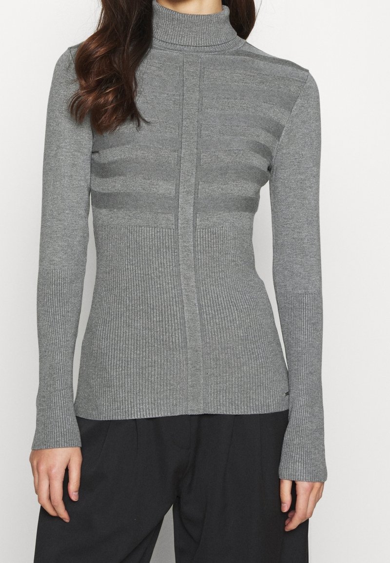 Femme portant un pull col roulé gris ajusté en maille côtelée avec de subtiles rayures horizontales et un pantalon noir ample, visible du cou jusqu'aux hanches.