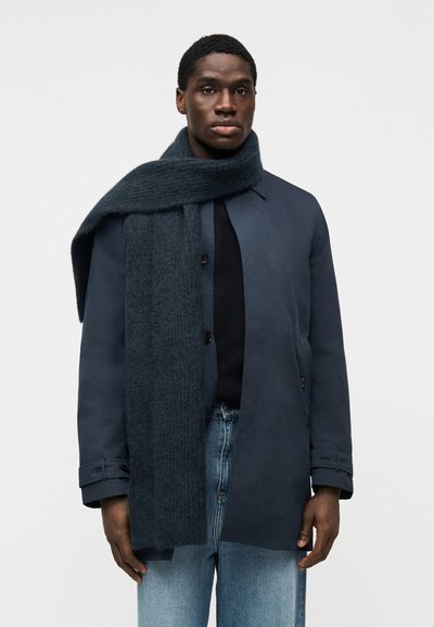 Manteau bleu marine avec un devant boutonné et un col; associé à une écharpe texturée foncée. Porté sur un haut noir et un jean bleu clair.