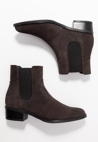 Donkerbruine suède enkellaarsjes met elastische zijpanelen, een lage hak van gestapeld materiaal en zichtbare stikselaccenten op een witte achtergrond.