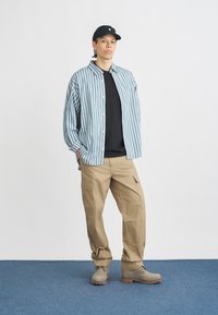 Mladý človek stojaci s rukami v vreckách, oblečený v čiernej čiapke, pruhovanej košeli v modro-bielej kombinácii, čiernom tričku, béžových cargo nohaviciach a čižmách.