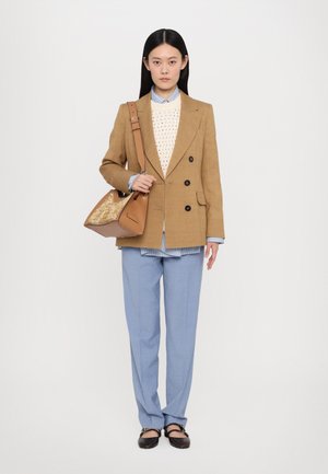 Tan dobbeltradet blazer over en hvid mønstret sweater, lyseblå skjorte og lyseblå bukser; holder en brun taske med en tekstureret front.