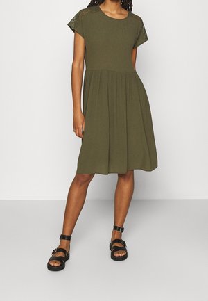 Frau in einem olivgrünen, knielangen Kleid mit Spitzenärmeln und schwarzen Open-Toe-Sandaletten mit Knöchelriemen, steht vor einem einfarbigen Hintergrund.