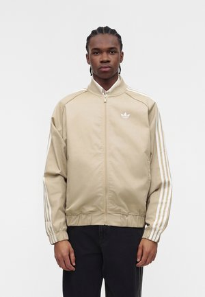 Mann mit geflochtenem Haar, der eine beige Adidas Jacke mit Reißverschluss und weißen Streifen an den Ärmeln sowie schwarze Hose trägt, steht vor einem einfarbigen Hintergrund.
