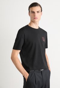 Zwarte katoenen t-shirt met een ronde hals, korte mouwen en een klein geborduurd rood "HUGO" logo op de linkerborst. Gladde textuur.