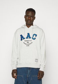 Pilkas hoodie su kengūrine kišenė ir virvute ant galvos. Yra mėlynos ir juodos kolegialinio stiliaus užrašys "AAC" ir "Adidas Athletic Club."