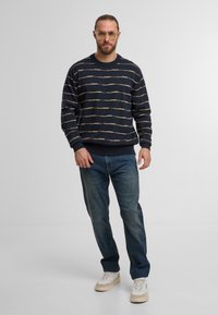 Maglione a righe navy con linee beige, polsini e orlo a costine; abbinato a jeans blu e sneakers bianche con accenti beige.