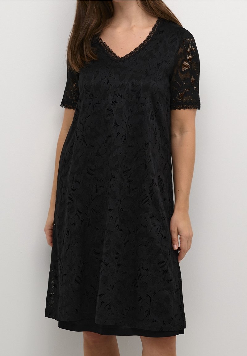 Femme portant une robe en dentelle noire longueur genou avec des manches courtes et un décolleté en V sur un fond clair uni.