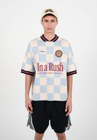 Ung mand iført blå og creme skjorte med tern og teksten "In a Rush", sorte shorts og beige kasket med teksten "Always Little Club".