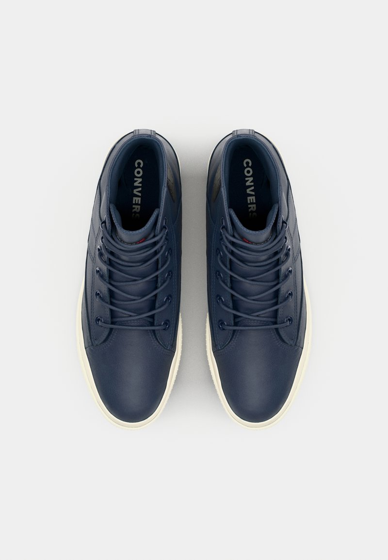 Marinblå lädersneakers med en jämn yta, vanligt snörningssystem och en beige gummisula. "CONVERSE" tryckt på insidan av tungan.