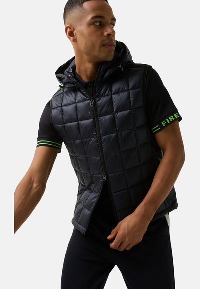 Schwarze gesteppte Weste mit Kapuze, ausgestattet mit einem vorderen Reißverschluss und grünen Akzenten. Getragen über einem kurzärmeligen schwarzen Shirt mit gemustertem Ärmel.