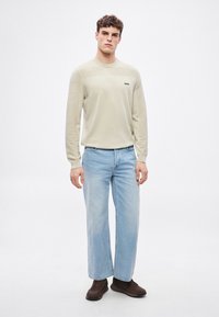 Világos bézs színű hosszú ujjú pulóver texturált vállrésszel és egy apró fekete "BOSS" logóval, laza szabású világoskék farmerral és barna tornacipővel párosítva.