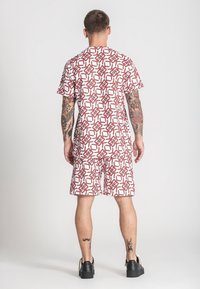 Košulja kratkih rukava sa uzorkom i odgovarajuće kratke hlače u bijeloj i crvenoj boji, s geometrijskim dizajnom. Tkanina izgleda lagano i glatko.