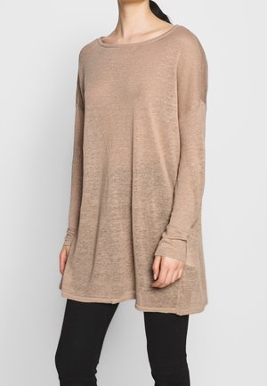 Kobieta ubrana w oversize'owy beżowy sweter-tunikę z długim rękawem i łódkowym dekoltem oraz czarne dopasowane spodnie na szarym tle.