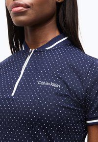 Κοντινό πλάνο ατόμου που φοράει μπλε σκούρο πόλο μπλούζα Calvin Klein με λευκές βούλες, άσπρο ριγέ γιακά και άκρες στα μανίκια.