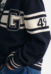 Granatowy dzianinowy kardigan z białymi paskami, z dużą literą "G" i "49" na rękawie. Paski ściągacza przy mankietach i kołnierzu dodają faktury.