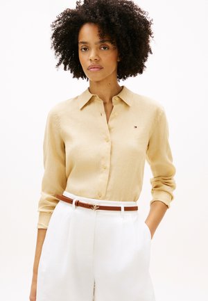 Kvinde med krøllet hår iført en beige skjorte med knapper, hvide bukser med høj talje og et brunt bælte med en guldbøjle, stående med den ene hånd i lommen.