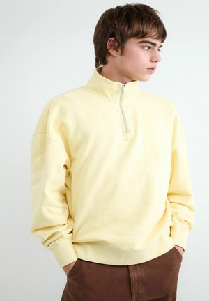 Mladý muž v bledožltej mikine so zipsom do štvrťky a hnedých nohaviciach, stojaci s rukami v vreckách, pozerajúc sa doprava na bielom pozadí.