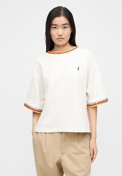 Biele bavlnené tričko so širokým, zaobleným výstrihom. Okolo výstrihu a rukávov má oranžové a červené lemovanie. Na hrudi vyšívané logo.