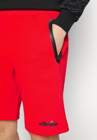 Persona que lleva pantalones cortos rojos brillantes con una mano en el bolsillo con cremallera lateral y el logo negro de ellesse cerca del dobladillo.