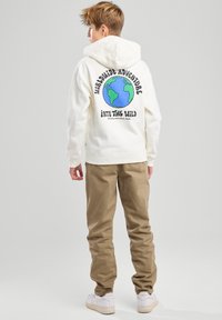 Weißer Hoodie mit einem bunten Erd-Design und Text auf der Rückseite, kombiniert mit khakifarbenen Hosen und weißen Sneakers. Glatte Stoffstruktur.