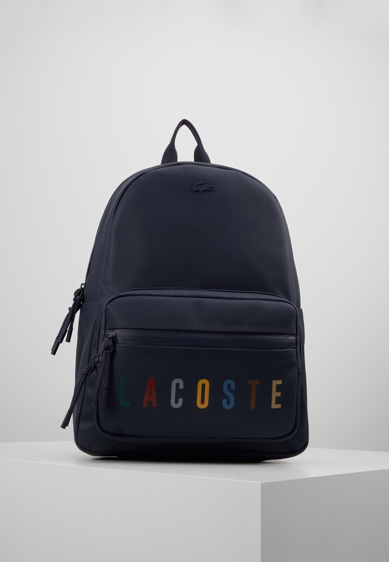 Navy blue reppure backpack with front zip pocket featuring multi-colored "LACOSTE" lettering and a small crocodile logo on top center.