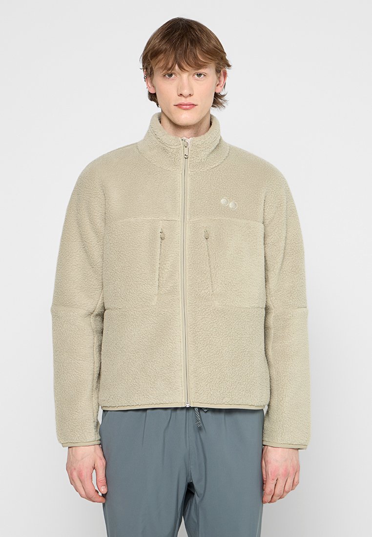 Veste en polaire beige avec un col haut, deux poches zippées à l'avant et un discret logo sur la poitrine gauche. Texture douce et coupe décontractée.