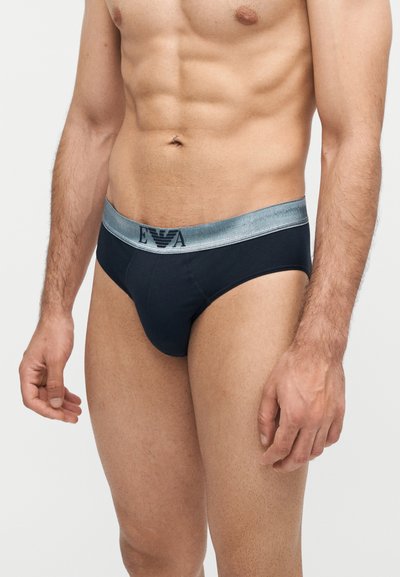 Άνδρας μοντέλο φοράει μαύρα εσώρουχα με γκρι λάστιχο μέσης με το λογότυπο "EVA", δείχνοντας τους τονισμένους κοιλιακούς και μέρος των χεριών του σε απλό φόντο.