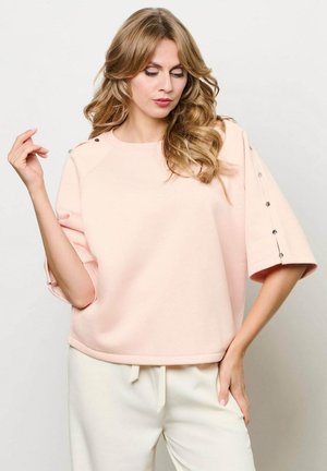 Vrouw met lang golvend haar draagt een lichtroze oversized top met knoopdetails op de mouwen en een witte broek met trekkoord, poserend tegen een effen achtergrond.