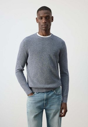 Hombre que lleva un jersey de punto gris claro sobre una camiseta blanca y vaqueros azules descoloridos, de pie con una mano en el bolsillo frente a un fondo liso.