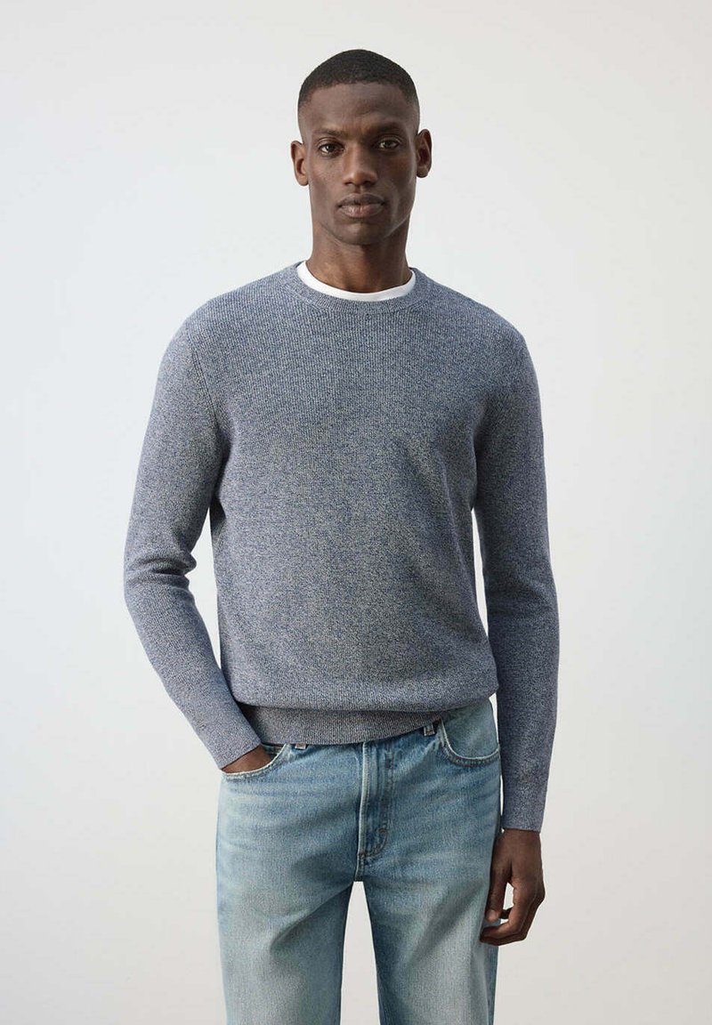Homme portant un pull en maille gris clair par-dessus un t-shirt blanc et un jean bleu délavé, debout avec une main dans la poche devant un fond uni.