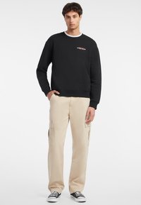 Svart hettegenser med ribbestrikket hals og brodert "GUESS JEANS"-logo, kombinert med beige cargo-bukser med sidelommer og løs passform.