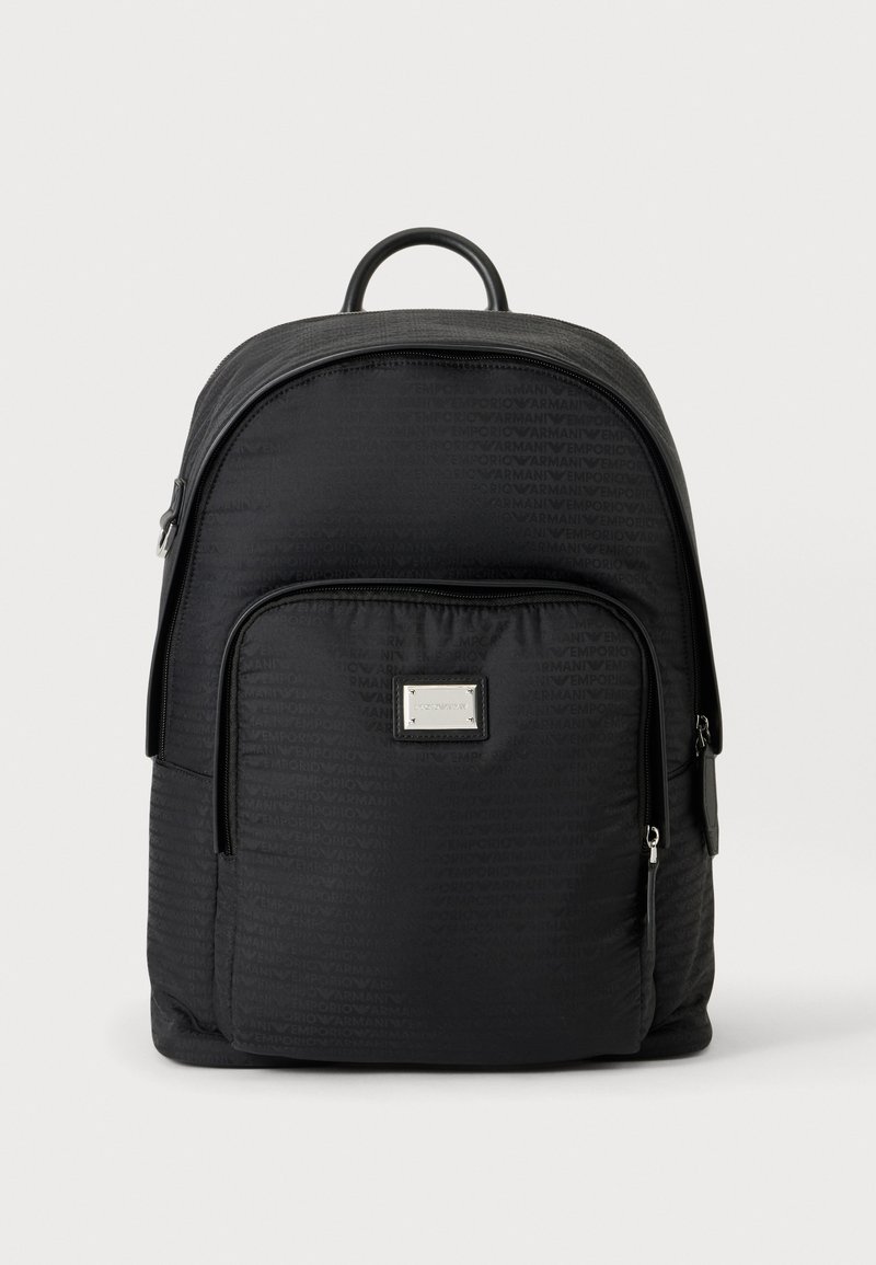 Sac à dos noir avec poche avant zippée, motif discret de logo, poignée supérieure et plaque rectangulaire argentée de la marque sur la poche avant.
