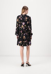 Abito nero floreale con maniche lunghe, vita cinchata e gonna fluida con motivi di fiori rosa e viola. Sono visibili tacchi alti neri con cinturini.