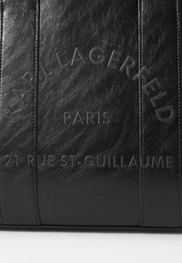Zwarte leren tas met reliëftekst: "KARL LAGERFELD PARIS 21 RUE ST-GUILLAUME." Beschikt over verticale naden en een gestructureerde afwerking.