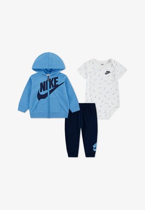 Ținuta formată din trei piese include un hanorac albastru cu fermoar, un body alb cu model și pantaloni bleumarin, toate având logo-uri Nike.