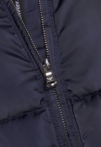 Marinblå pufferjacka som visar en nära bild av en silverfärgad metalldragkedja och placket, med kontrasterande sömmar och en slät, glänsande tygkänsla.