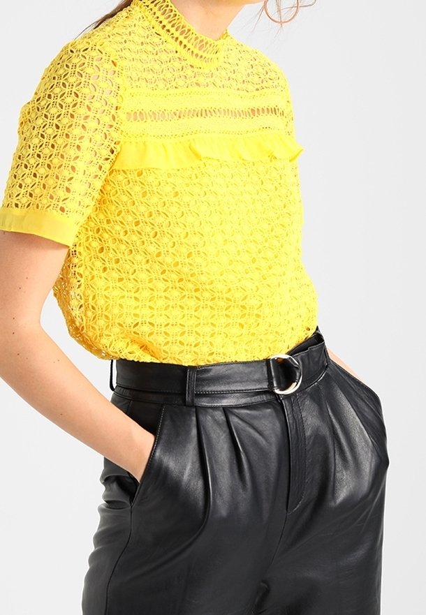 Femme portant un chemisier en dentelle jaune vif à manches courtes rentré dans un pantalon en cuir noir taille haute avec ceinture, les mains dans les poches.