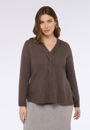 Kobieta z włosami sięgającymi do ramion, w brązowym swetrze z dzianiny i złotych kolczykach w kształcie kół, nosząca jasnoszary spódnicę, na jednolitym tle.